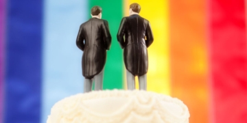 This image shows the top of a traditional wedding cake, with two men in tuxedos and tails as the wedding cake topper and with a rainbow "pride" flag in the background.
