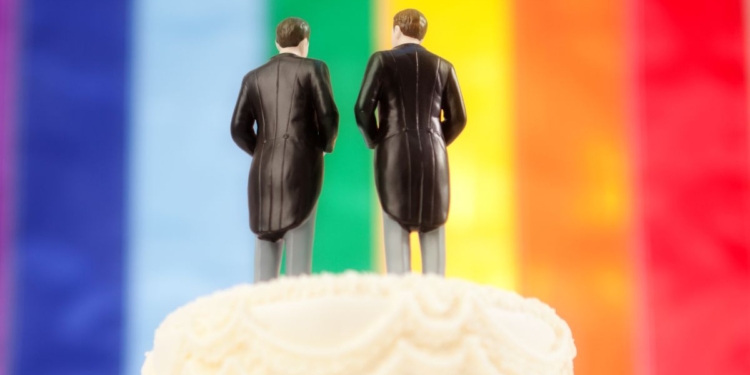 This image shows the top of a traditional wedding cake, with two men in tuxedos and tails as the wedding cake topper and with a rainbow "pride" flag in the background.